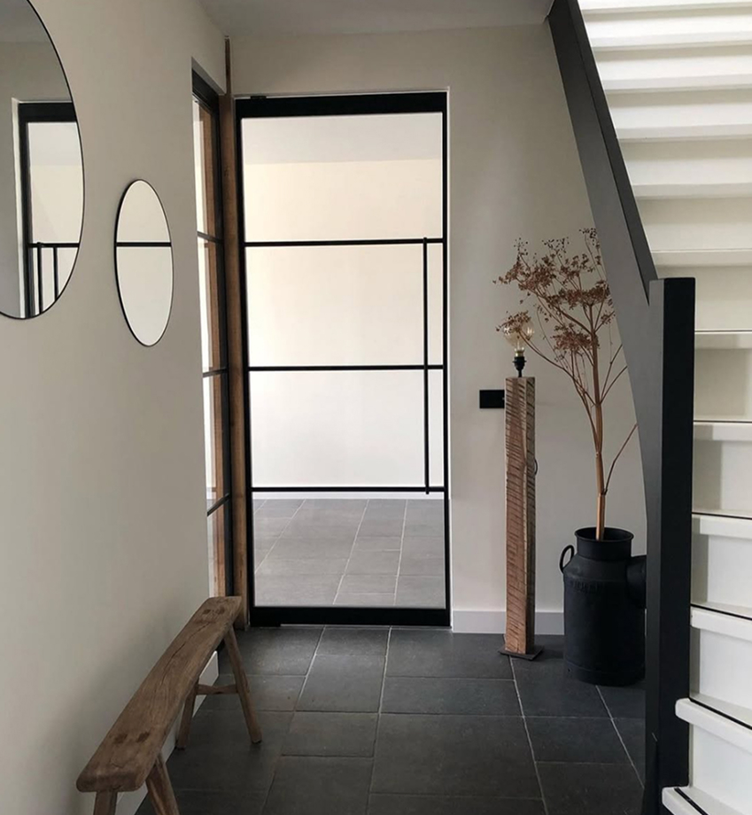 Flur mit schwarzem Metallrahmentür, zwei runden Spiegeln an der Wand, Holzbänkchen und Treppe mit weißen Stufen. Flur mit schwarzem Metallrahmentür, zwei runden Spiegeln an der Wand, Holzbänkchen und Treppe mit weißen Stufen.