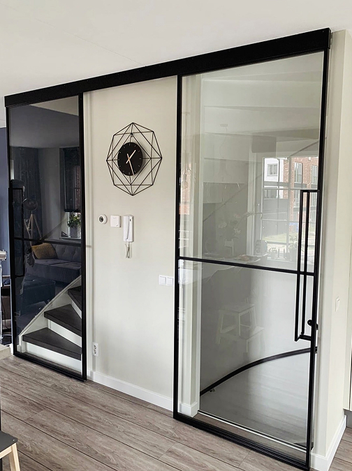 Moderner Wohnraum mit schwarzem Metallrahmen, Glastür mit Griff, geometrischer Wanduhr und Treppe im Hintergrund. Moderner Wohnraum mit schwarzem Metallrahmen, Glastür mit Griff, geometrischer Wanduhr und Treppe im Hintergrund.