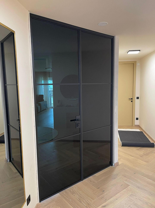 Schwarze, rahmenlose Glasschiebetür in einem hellen Flur mit Parkettboden und beige lackierter Zimmertür im Hintergrund. Schwarze, rahmenlose Glasschiebetür in einem hellen Flur mit Parkettboden und beige lackierter Zimmertür im Hintergrund.